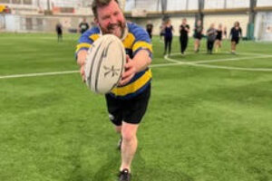 masc individual with beard withing striped shirt holding rugby ball and smiling